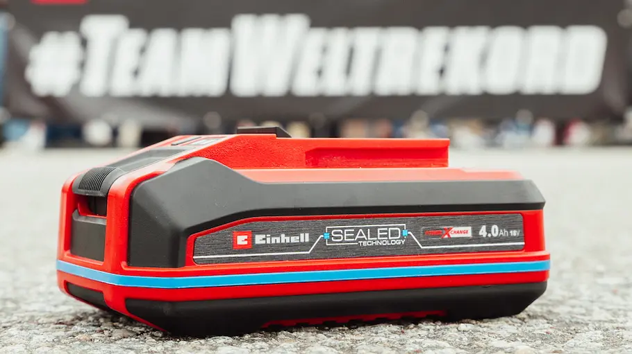 Ein roter Einhell 18V Sealed Akku liegt auf dem Boden vor einem Banner mit der Aufschrift #TeamWeltrekord, im Hintergrund sind unscharfe Personen zu sehen.