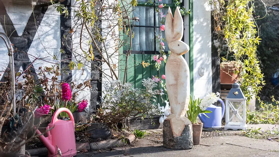 Ein aus Holz geschnitzter Osterhase steht als Gartendekoration auf einer Terrasse zwischen Frühlingsblumen, Laterne und Blumentöpfen vor einem Haus.