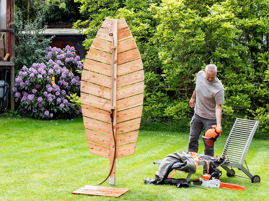 Die fertige Gartendusche im Garten Leaf-förmige Gartendusche aus hellen Holzlatten steht auf einer Rasenfläche; im Hintergrund ein Mann mit Schutzhelm neben einem Holzliegestuhl, Arbeitskleidung und einer Kettensäge, dahinter blühende Rhododendren und grüne Hecken.