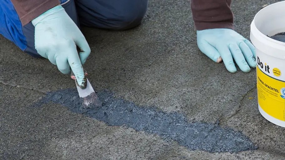 Mit einem Spachtel wird schwarze Bitumendichtmasse in einen Spalt zwischen zwei Dachpappen eingepresst