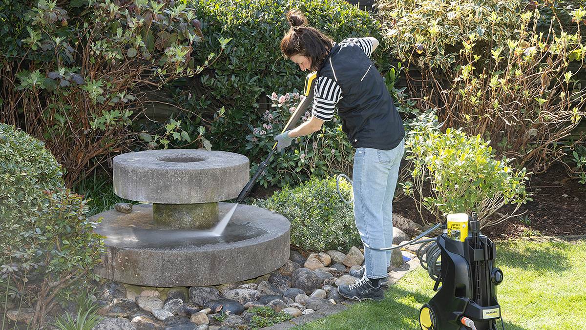 Eine Frau spritzt einen steinernen Brunnen im Garten mit einem Hochdruckreiniger ab, um ihn zu säubern.