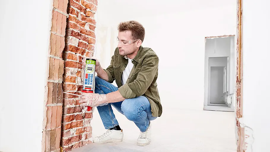 Bauschaum in Mauerfugen Ein Mann mit Schutzbrille und Handschuhen sprüht Bauschaum in eine Lücke zwischen Ziegelwand und Türrahmen.