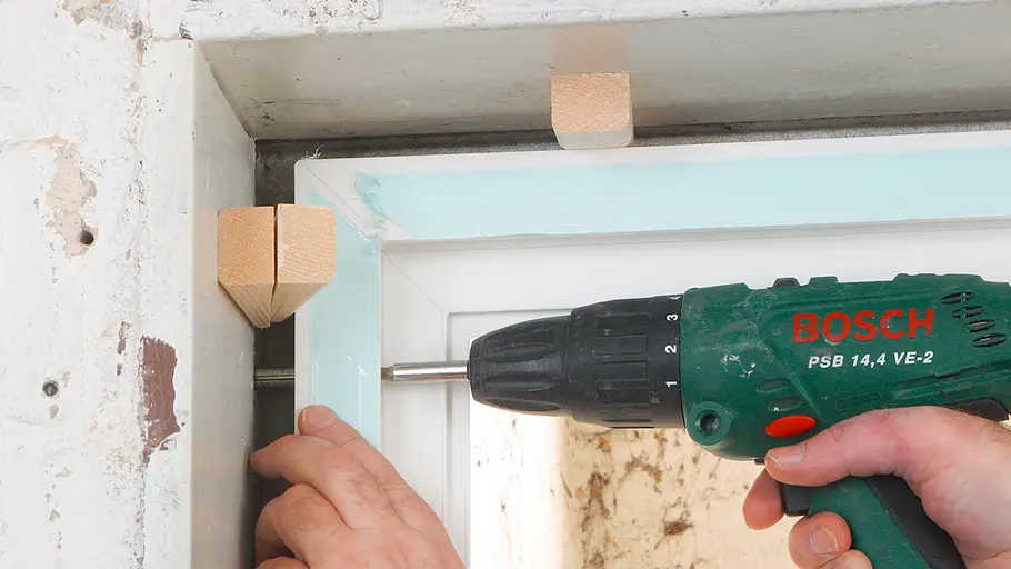 Fenster verkeilen Ein Akkuschrauber wird verwendet, um einen Fensterrahmen mithilfe von Holzkeilen und Dübeln in einer Wand zu befestigen.