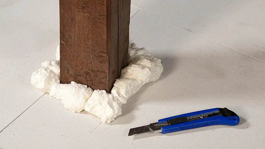 Überstände abschneiden Weiße Bauschaummasse umschließt den Fuß eines Holzbalkens auf hellen Fliesen, daneben liegt ein blaues Cuttermesser.