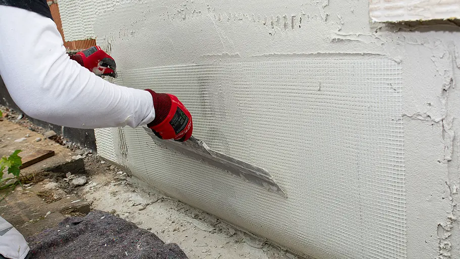 Dämmen und verputzen Eine Person mit roten Handschuhen zieht frischen Putz mit einer Glättkelle über ein eingelegtes Armierungsgewebe an der Hauswand.