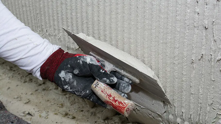 Dämmen und verputzen Eine Hand mit Handschuh verteilt frischen Kleber mit einer Zahnkelle in Rillenstruktur auf einer Wand mit Dämmplatten.