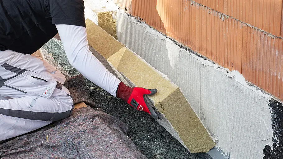 Dämmen und verputzen Eine Person im weißen Arbeitsanzug drückt eine Dämmplatte gegen einen mit Kleber bestrichenen Sockel einer Ziegelhauswand.
