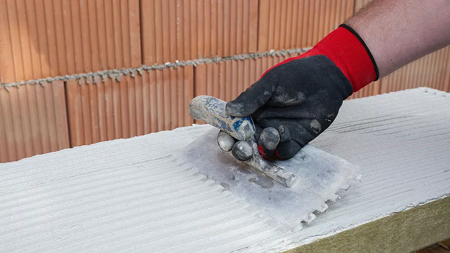 Dämmen und verputzen Eine Hand mit schwarzem und rotem Handschuh verteilt weißen Mörtel mit einer Zahnkelle auf einer Dämmplatte vor Ziegelwand.