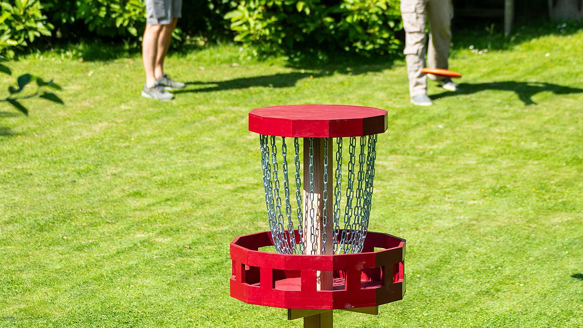 Ein Discgolf-Korb steht auf einer grünen Wiese. Im Hintergrund sind Bäume sichtbar und zwei paar Beine von den Discgolf-Spielern.