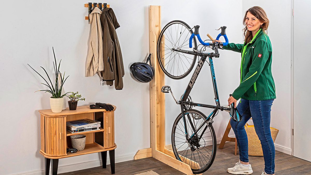Eine sportlich gekleidete junge Frau stellt ihr Fahrrad in einen Fahrradständer. Der Fahrradständer befindet sich im Flur einer Wohnung, links daneben eine Garderobe mit Jacken und eine kleine Kommode. Der Fahrradständer ist aus Holz und L-förmig, wobei die lange Seite an der Wand befestigt ist und die kurze Seite am Boden. Das L besteht aus einer langen Laufschiene, in die das Fahrrad gestellt wird. Das Vorderrad in die Laufschiene an der Wand, das Hinterrad in der Laufschiene auf dem Boden. An einem Haken an der linken Seite des Fahrradständers hängt ein Fahrradhelm.
