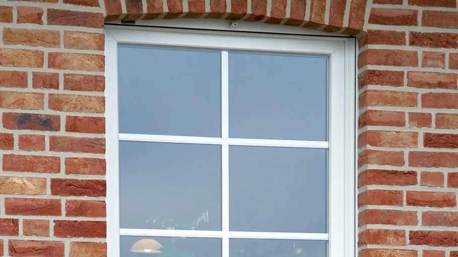 Fenster auswählen Ein weißes Fenster mit Sprossen ist in eine rote Backsteinfassade mit leichtem Rundbogen über dem Rahmen eingebaut.