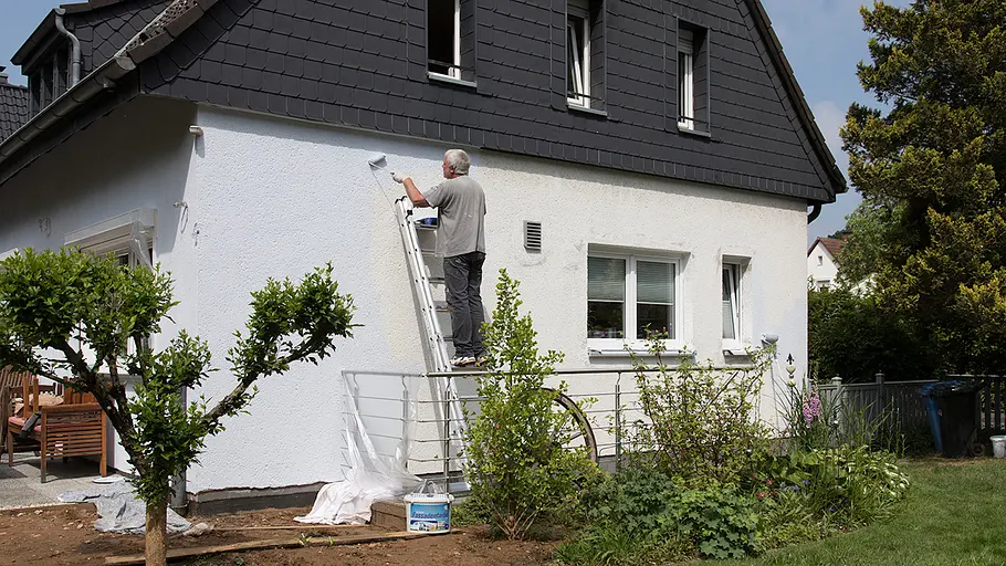 Fassade streichen Ein Mann steht auf einer Leiter und streicht mit einer Rolle helle Fassadenfarbe an die Außenwand eines Einfamilienhauses.