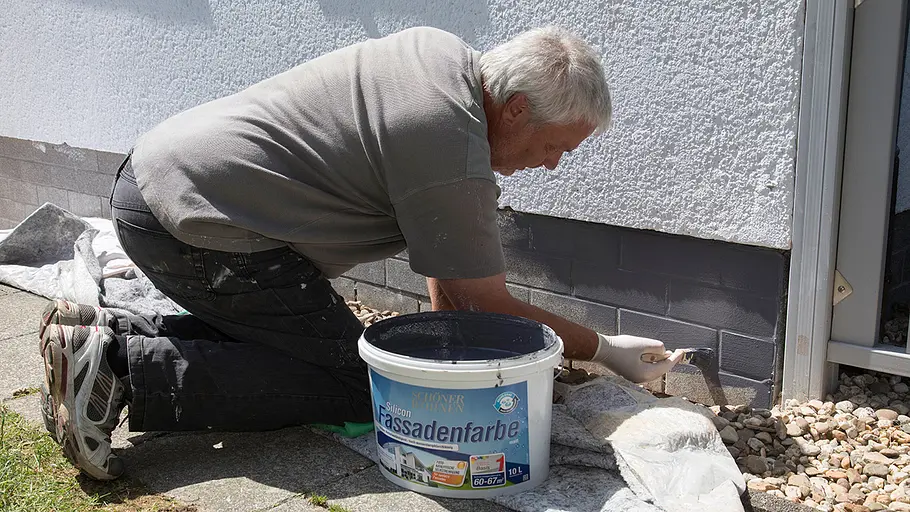 Fassade streichen Ein Mann kniet vor dem Haus und streicht mit Pinsel dunkle Fassadenfarbe auf den unteren Bereich der Hauswand.