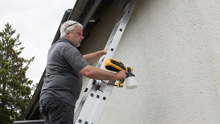 Fassade streichen Ein Mann steht auf einer Leiter und trägt mit einer Farbspritzpistole Farbe auf die Hausfassade auf.