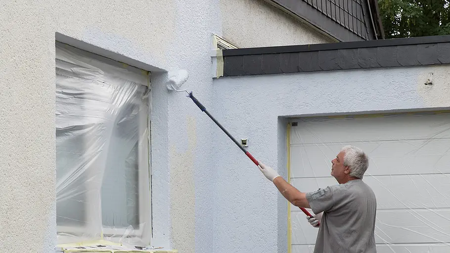 Fassade streichen Ein Mann mit weißem Haar streicht mit einer Teleskoprolle weiße Farbe auf eine Hausfassade, Fenster und Tür sind abgeklebt.