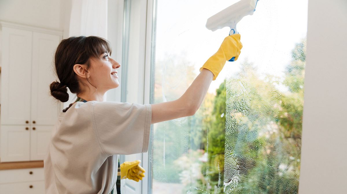 Ratgeber: Fenster streifenfrei putzen eine junge Frau mit hochgebundenen braunen Haaren putzt mit einem Fensterwischer lächelnd ein Fenster, zum Schutz vor der Seifenlauge trägt sie leuchtend-gelbe Gummihandschuhe