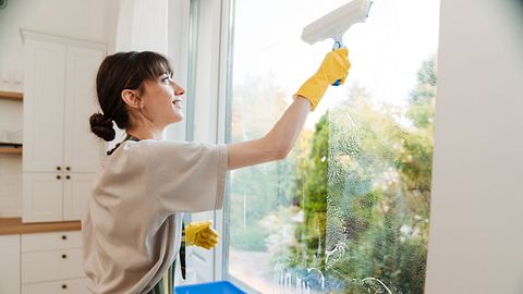 eine junge Frau mit hochgebundenen braunen Haaren putzt mit einem Fensterwischer lächelnd ein Fenster, zum Schutz vor der Seifenlauge trägt sie leuchtend-gelbe Gummihandschuhe - Foto: AdobeStock / Drobot Dean