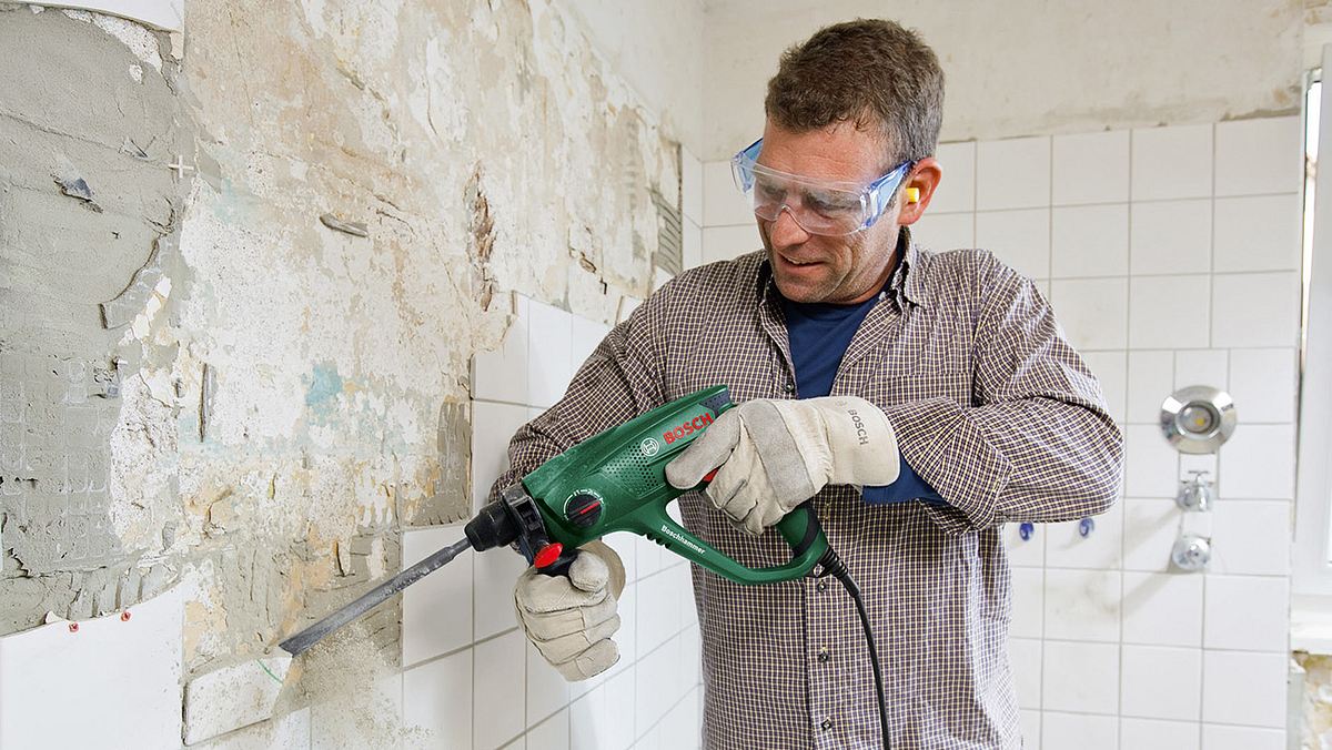 Mann entfernt Fliesen an der Wand mit einem Bohrhammer mit Meißelaufsatz.