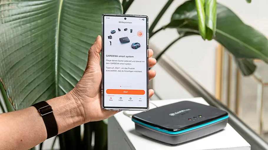 Gardena Smart Water Control Eine Person hält ein Smartphone mit geöffneter Gardena-App, daneben steht die Gardena Smart Water Control Basisstation.