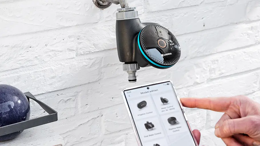 Gardena Smart Water Control Ein Gardena Smart Water Control ist am Wasserhahn installiert, daneben bedient eine Hand ein Smartphone mit passender App.