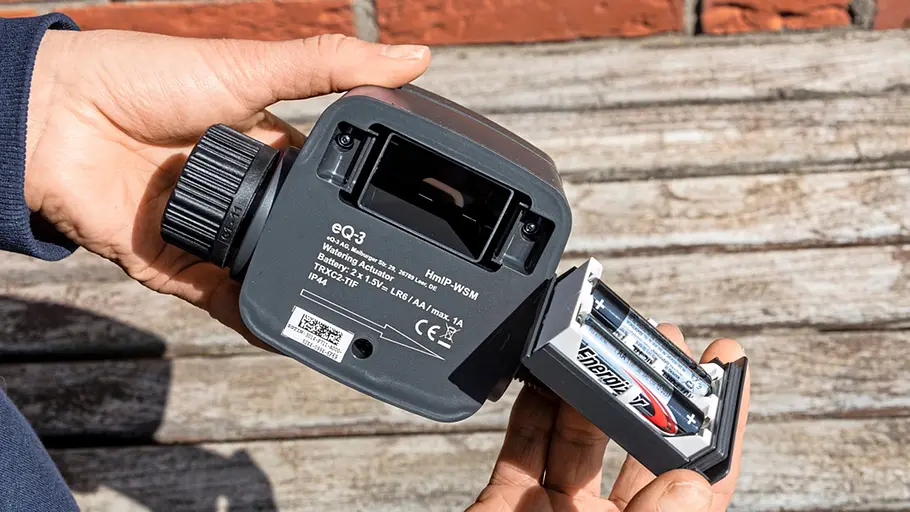 Homematic IP Bewässerungsaktor Eine Person hält einen Homematic IP Bewässerungsaktor und setzt frische Batterien in das geöffnete Batteriefach ein.