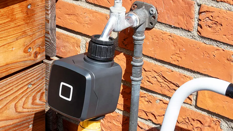 Homematic IP Bewässerungsaktor Ein Homematic IP Bewässerungsaktor ist an einem silbernen Wasserhahn vor einer roten Backsteinwand angeschlossen.
