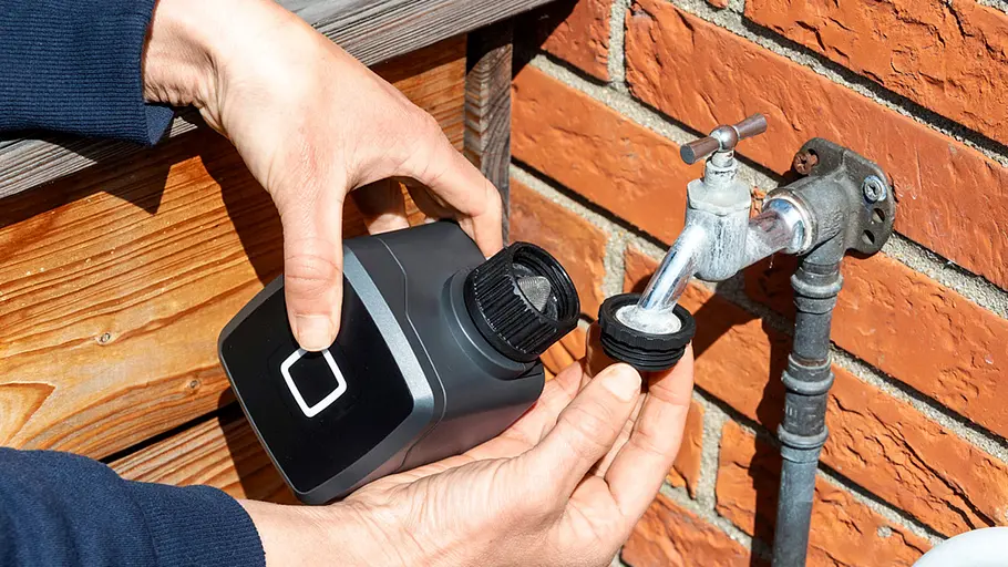 Homematic IP Bewässerungsaktor Eine Person montiert einen Homematic IP Bewässerungsaktor an einem Wasserhahn vor einer roten Backsteinwand.