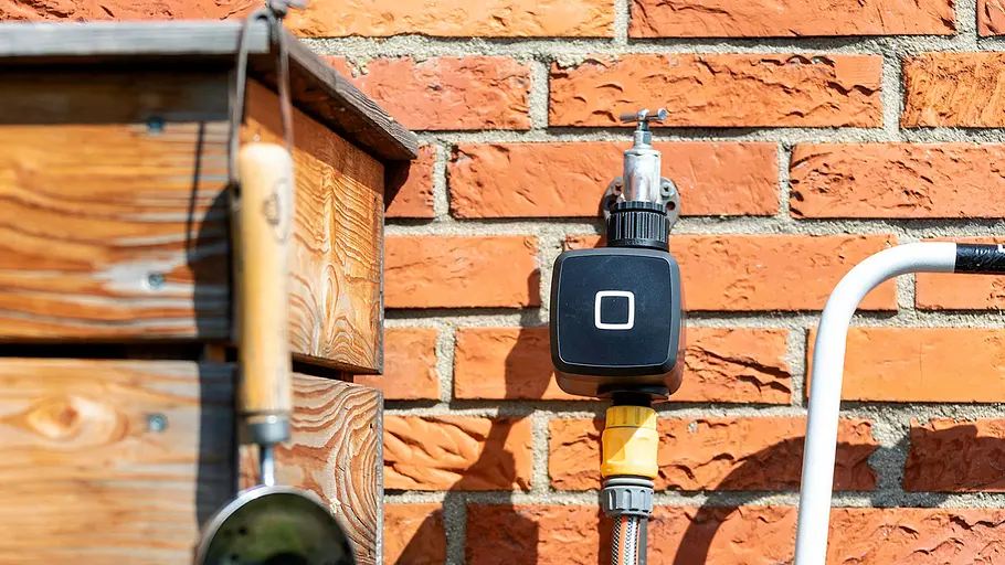 Homematic IP Bewässerungsaktor Ein Homematic IP Bewässerungsaktor ist an einem Wasserhahn vor einer roten Backsteinwand angeschlossen.
