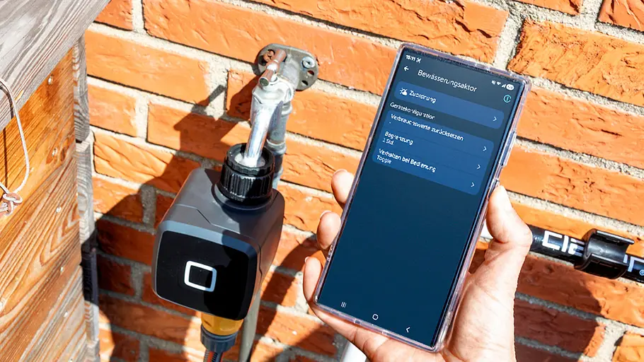 Homematic IP Bewässerungsaktor Ein Homematic IP Bewässerungsaktor ist am Wasserhahn installiert, daneben steuert eine Hand das Gerät per Smartphone-App.