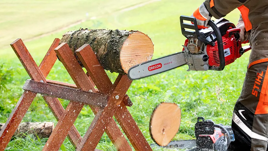Schnelligkeitstest Kettensägen-Vergleich Eine Person mit Schutzkleidung schneidet mit einer roten Motorsäge ein Holzstück von einem Stamm auf einem Sägebock ab.