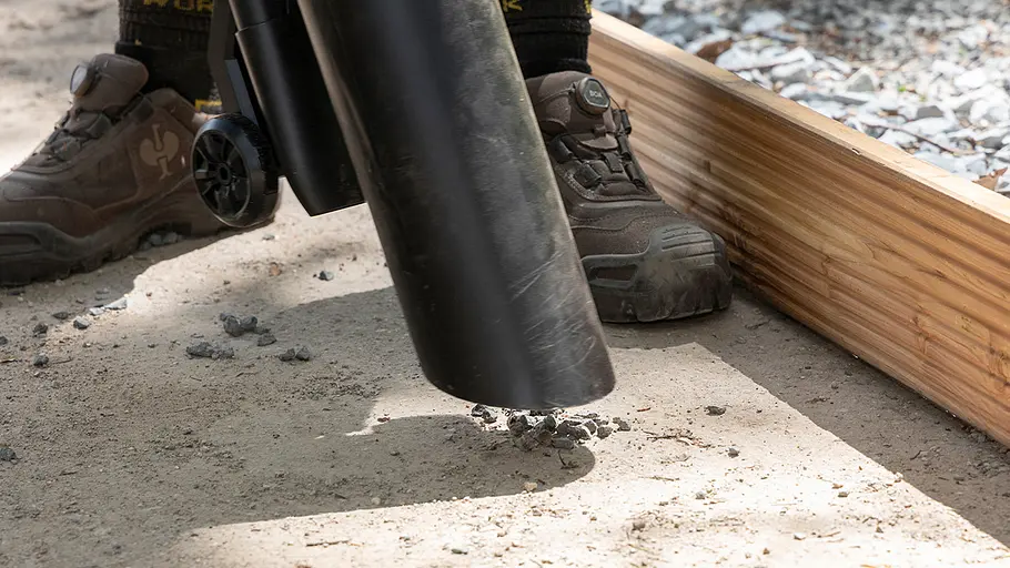 Die Düse eines Laubbläsers saugt kleine Steine nahe eines Holzrahmens auf, zwei Füße mit festen Schuhen daneben.