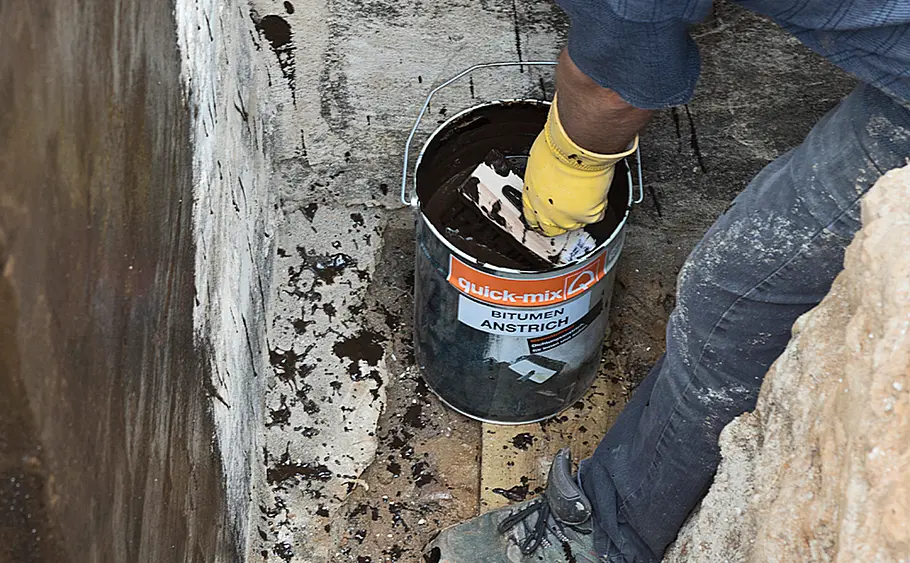 Bitumenanstrich-Eimer vor Hauswand mit Person Eine Person mit gelbem Handschuh taucht einen Quast in einen offenen Eimer Bitumenanstrich vor einer Hauswand.