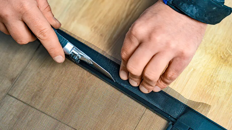 Zwei Hände schneiden mit einem silbernen Cuttermesser das überstehende Insektengitter am schwarzen Rahmen ab.