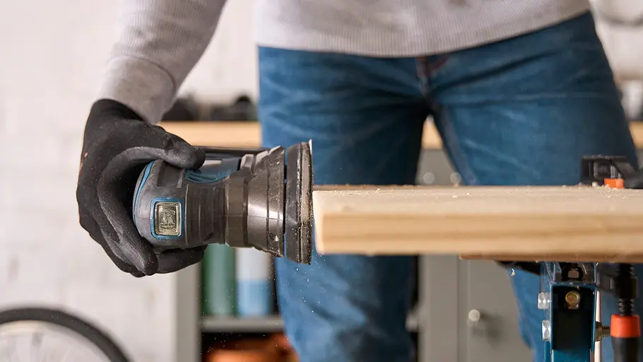 Matschküche: Schnittkanten glätten Eine Person hält einen elektrischen Schleifer und glättet die Schnittkante einer Holzplatte, die auf einer Werkbank liegt.
