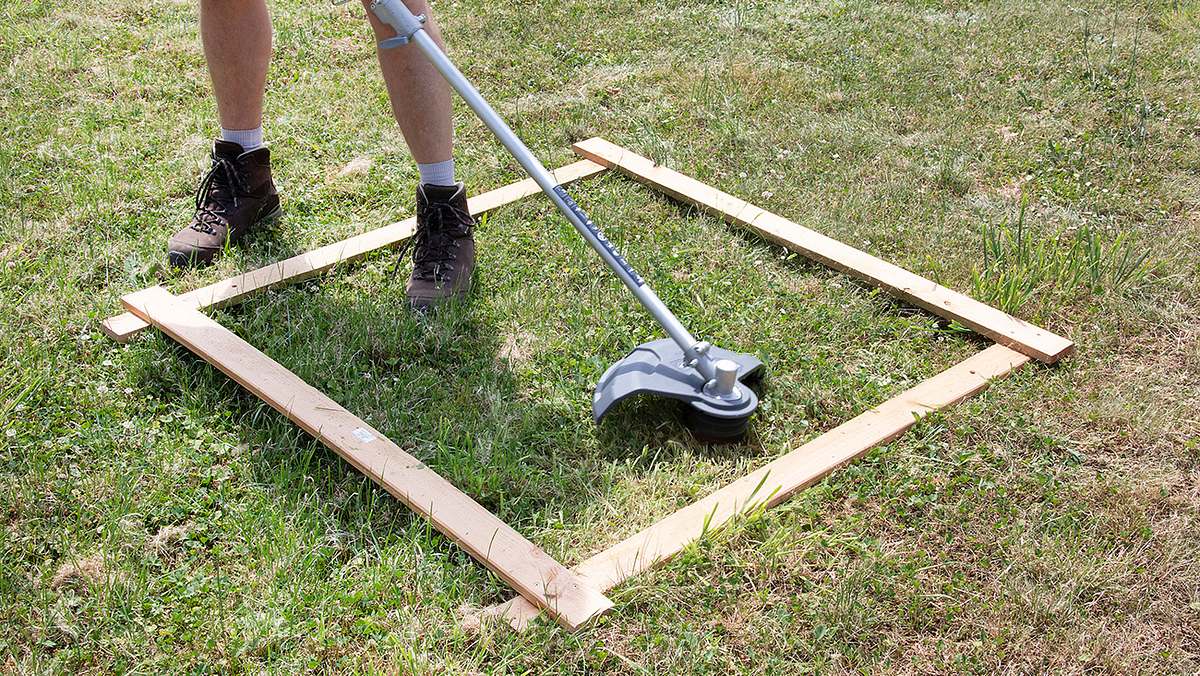 Ein Akku-Rasentrimmer wird von einer Person mit Arbeitsstiefeln innerhalb eines Holzrahmens auf einer Wiese getestet.
