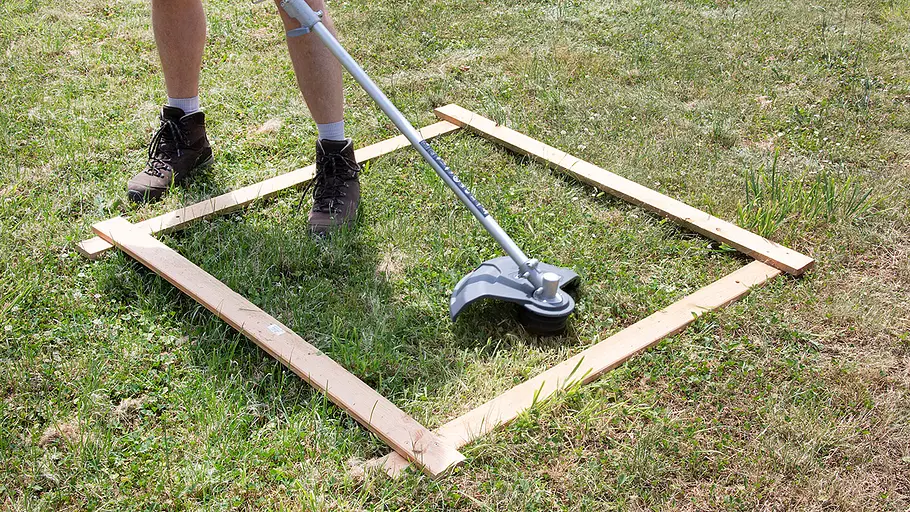 Ein Akku-Rasentrimmer wird von einer Person mit Arbeitsstiefeln innerhalb eines Holzrahmens auf einer Wiese getestet.