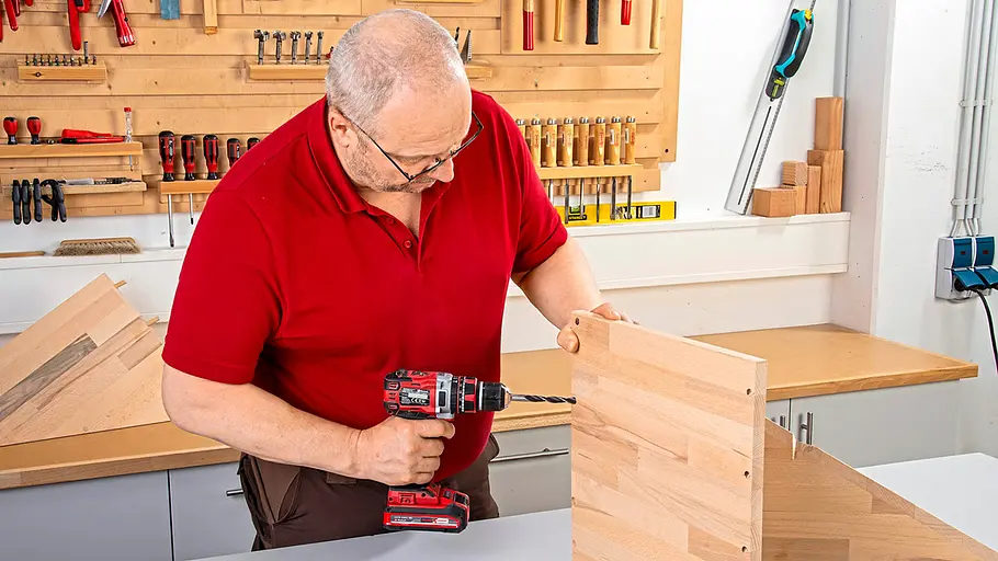 Verdrehter Hocker Ein Mann bohrt mit einer Akku-Bohrmaschine Löcher in eine aufgestellte Holzplatte in einer Werkstatt.