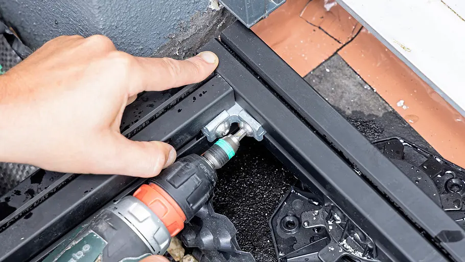 Terrasse gestalten Ein Akkuschrauber fixiert einen Metallverbinder zwischen zwei schwarzen Profilen auf Unterkonstruktion beim Terrassenbau.