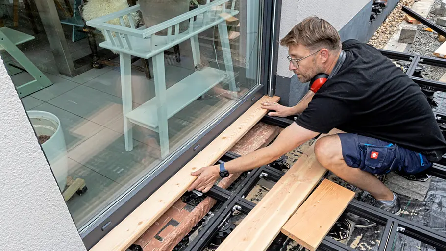 Terrasse gestalten Ein Mann legt ein zugeschnittenes Holzbrett an der Terrassentür passgenau auf die Unterkonstruktion der Terrasse.