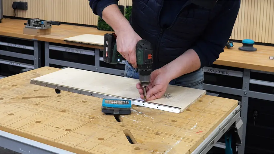 Paketbox Eine Person schraubt mit einem Akkuschrauber eine Metallschiene an eine Holzplatte auf einer Werkbank.