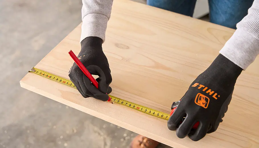 Matschküche: Platten zuschneiden Eine Person trägt schwarze STIHL-Handschuhe und misst mit Maßband und Bleistift eine Holzplatte zum Zuschneiden ab.