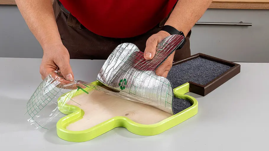 Kratzmatte anbringen Eine Person legt eine zugeschnittene, selbstklebende Folie mit Teppich auf das grün lackierte Kaktus-Kratzbrett.