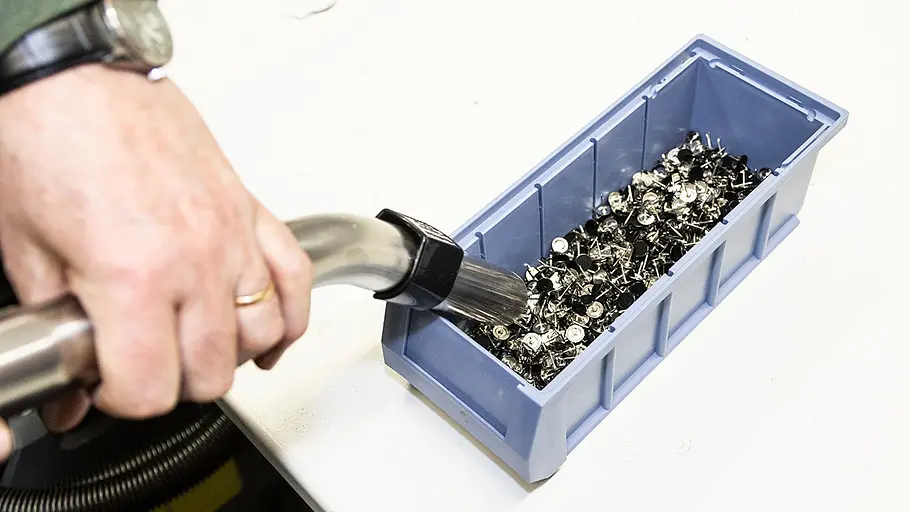 Werkstattsauger Härtetest Eine Hand hält den Schlauch eines Werkstattsaugers und saugt Metallteile aus einer blauen Kunststoffkiste auf einem Tisch.