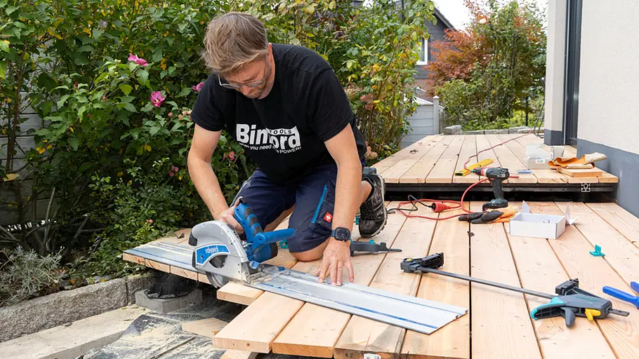 Terrasse gestalten Ein Mann schneidet mit einer Handkreissäge und Führungsschiene die überstehenden Terrassendielen exakt ab.