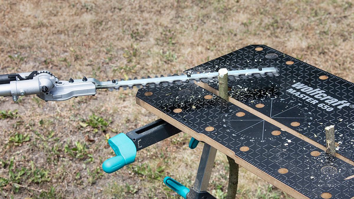 Eine Akku-Heckenschere schneidet einen dicken Ast auf einem Arbeitsbock im Freien, Holzspäne sind zu sehen.