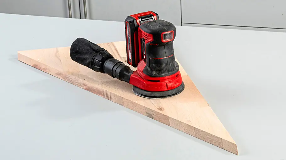 Verdrehter Hocker Ein Dreieck-Holzbrett liegt auf einer Werkbank, darauf steht ein roter kabelloser Exzenterschleifer mit Staubfang.