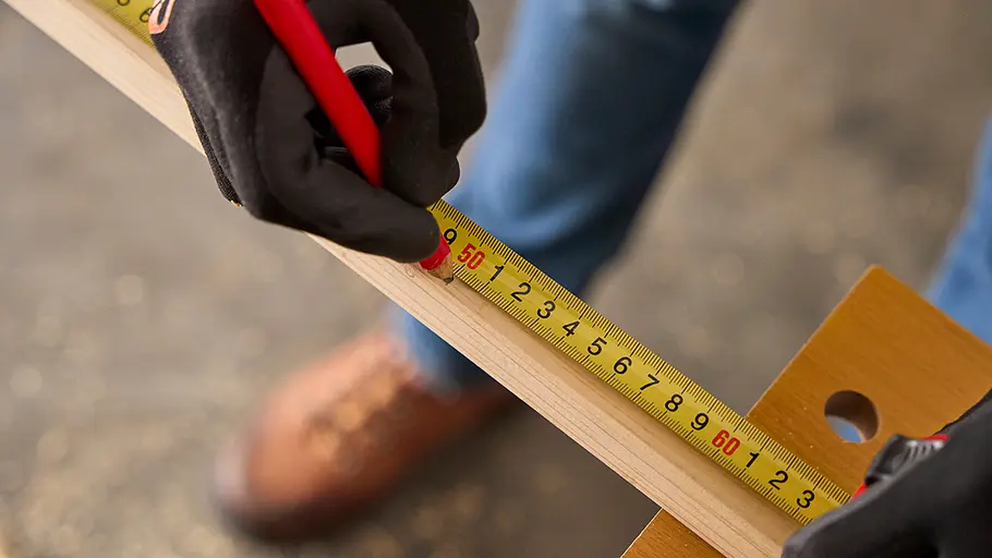 Eine Person mit schwarzen Handschuhen markiert mit einem Stift und Maßband ein Maß auf einem Holzstab.