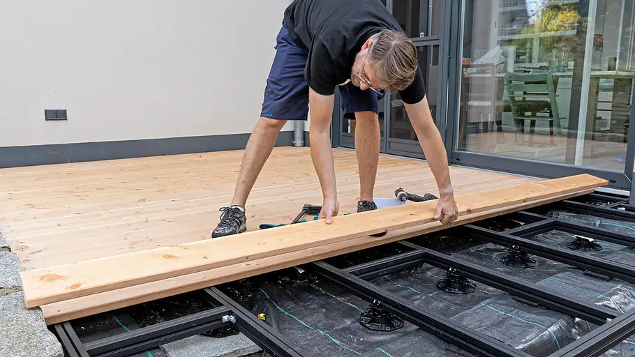 Terrasse gestalten Ein Mann legt ein langes Holzbrett auf die Unterkonstruktion, um die Terrasse weiter zu beplanken.
