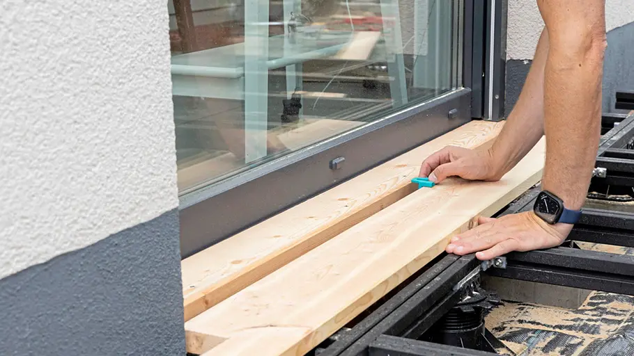 Terrasse gestalten Eine Person markiert mit einem Bleistift eine Schnittlinie auf einer Holzterrassendiele direkt an der Terrassentür.