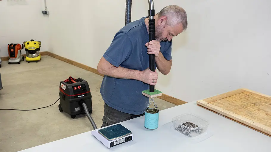 Ansaugstärke-Test Ein Mann prüft mit einem Werkstattsauger und einer Dose auf einem Tisch die Saugkraft, daneben liegen Metallkugeln und eine Waage.